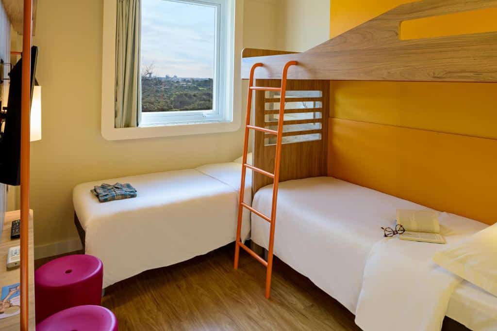 Quarto do ibis budget Foz do Iguaçu que mostra uma beliche e do lado uma cama de solteiro do lado de uma janela também. No quarto tem dois banquinhos e uma tv na parede.