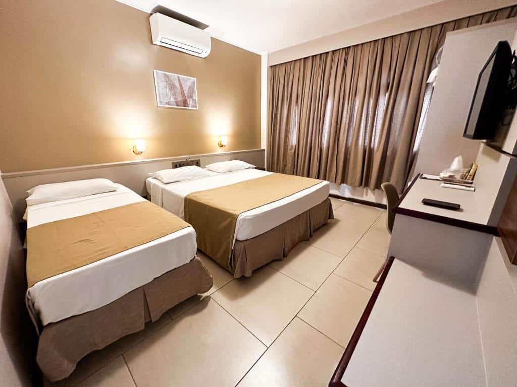 Quarto do San Rafael Confort com uma cama de casal e do lado uma cama de solteiro. Na parede atrás das camas tem um quadro e um ar-condicionado e na parede do lado tem uma janela com cortina. Na frente das camas tem uma escrivaninha com cadeira e uma tv na parede.