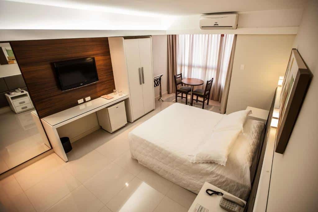 Quarto do Rafain Palace Hotel & Convention Center com uma cama de casal do lado direito com uma mesa de cabeceira de cada lado. Em frente da cama tem uma mesa, uma tv na parede, um frigobar, um guarda-roupa e do lado uma mesa redonda com duas cadeiras, um ar-condicionado e uma janela com cortina.