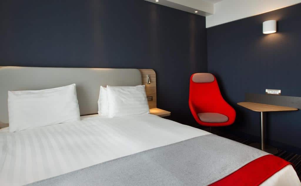 Foto de um quarto no Holiday Inn Express Geneva Airport, para ilustrar o post de hotéis em Genebra. A cama é box de casal, e vemos ela de frente, ocupando a maior parte esquerda da foto. Na direita, há uma poltrona vermelha com mesa. As paredes do quarto são azul escuro.