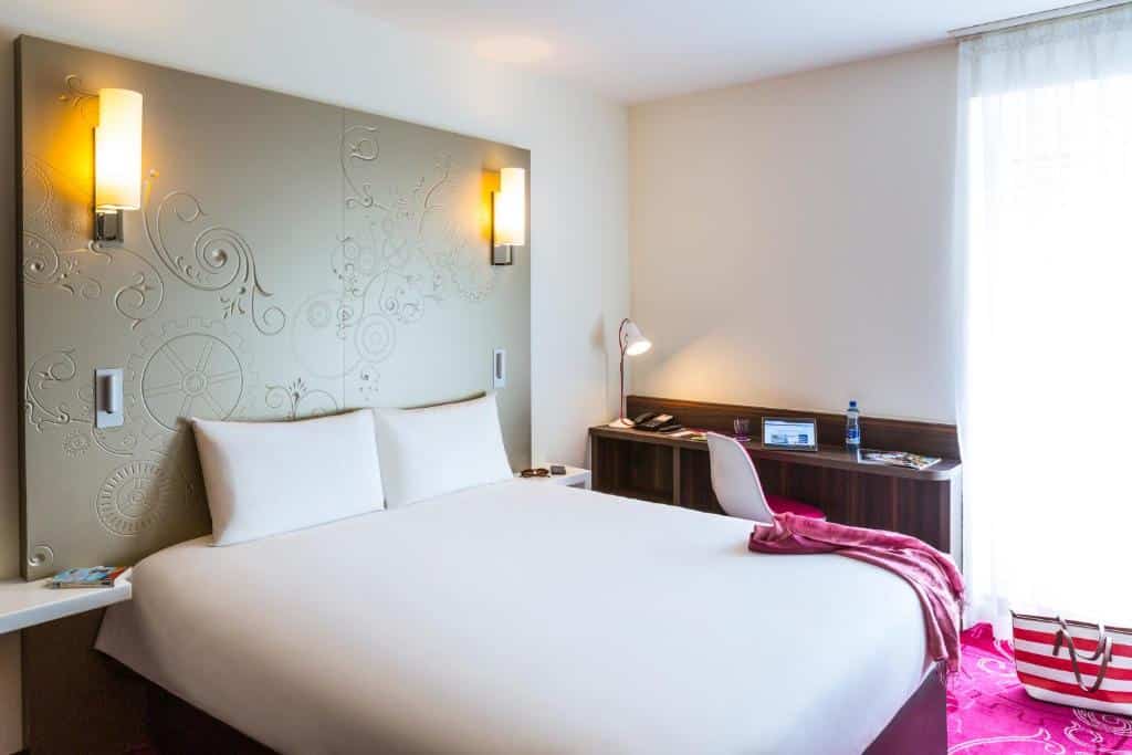 Foto de um quarto no VISIONAPARTMENTS Geneva Mont-Blanc, para ilustrar o post de hotéis em Genebra. A cama box de casal está encostada em uma cabeceira cinza de ferro, na esquerda do quarto. No lado direto da cama, encostada na parede do fundo, há uma escrivaninha de madeira com abajur, telefone, água e um tablet, acompanhada de uma cadeira. A janela está do lado, na direita. Uma bolsa listrada está no chão, perto da janela.