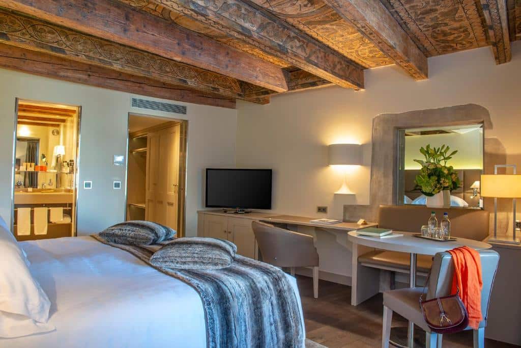 Foto de um quarto do Les Armures, para ilustrar o post de hotéis em Genebra. Uma cama de casal está no canto inferior esquerdo da foto, de costas para a imagem. Na frente da cama, há uma escrivaninha com TV em cima, abajur e duas cadeiras. Um espelho também está fixo na parede. No lado esquerdo do quarto, do lado da escrivaninha está a porta da suíte e outra porta para o banheiro.