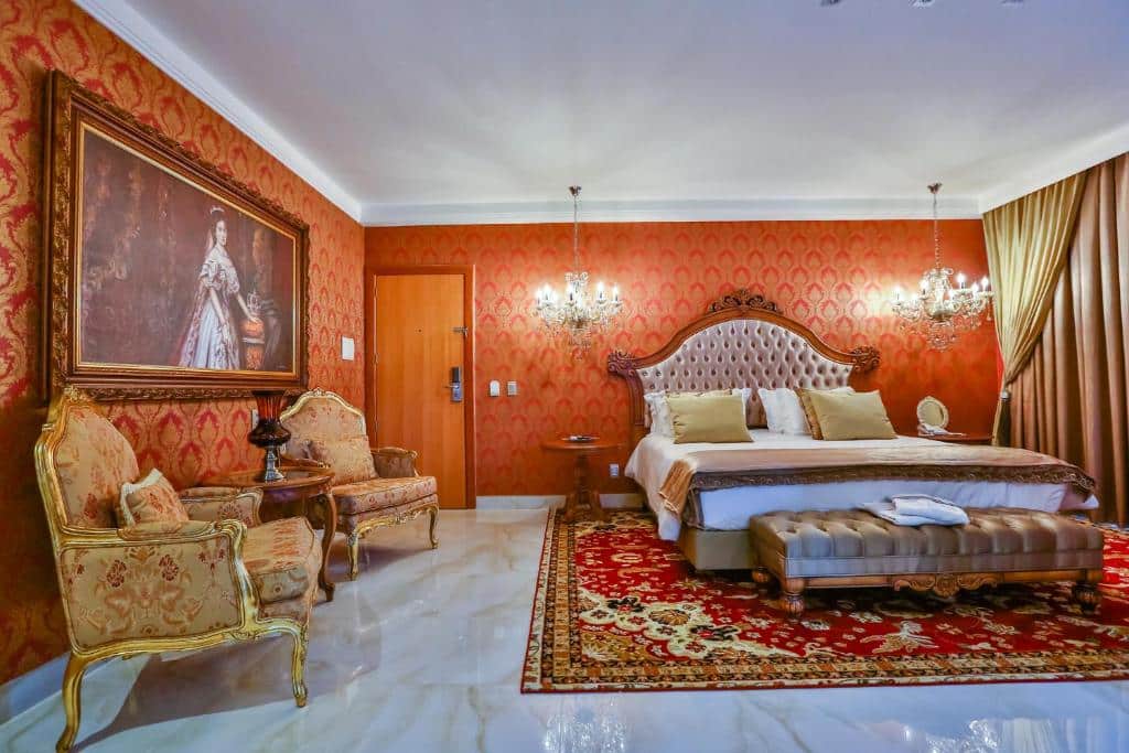 Quarto de hotel luxuoso com decoração temática inspirada na cidade de Viena do século XIX. No lado esquerdo do quarto estão duas poltronas grandes e um quadro antigo a cima, no lado direito está uma cama king size com cabeceira e lustres ao lado da cama, em baixo da cama há um tapete e as paredes são revestidas com papel decorativo. Imagem para ilustrar o post hotéis em Goiânia.