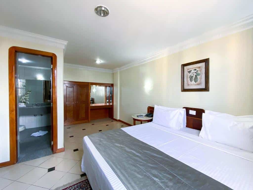 Quarto simples com cama de casal grande com colcha branca e cinza, quadro decorativo em cima da cama, porta em frente a cama para o banheiro e armário de madeira no fundo do quarto. Imagem para ilustrar o post hotéis em Goiânia.