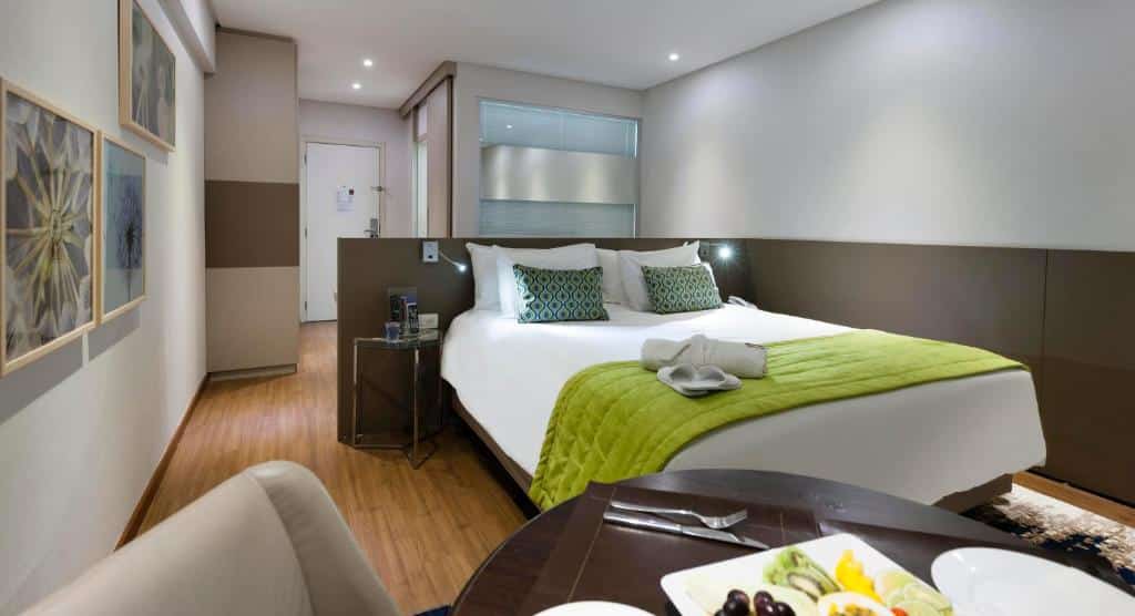 Quarto de hotel espaçoso com paredes brancas, cama de casal com colcha branca e verde, mesinha de vidro ao lado e mesa com café da manhã a frente da cama. Imagem para ilustrar o post hotéis em Goiânia.