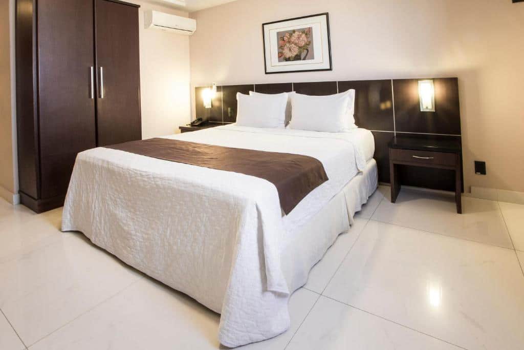 Quarto com cama de casal com colcha branca e detalhe em marrom, cabeceira de madeira, armário de madeira no lado esquerdo da cama, ar-condicionado, quadro decorativo em cima de cama e abajour ao lado. Imagem para ilustrar o post hotéis em Goiânia.