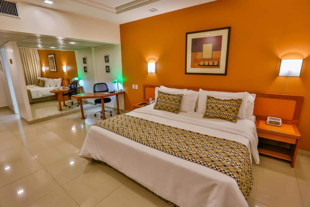 Quarto amplo com paredes brancas e laranja, cama de casal com colcha branca e detalhes estampados, quadro decorativo em cima da cama, abajour nos lados, mesa, cadeira e espelho grande no lado esquerdo do quarto. Imagem para ilustrar o post hotéis em Goiânia.