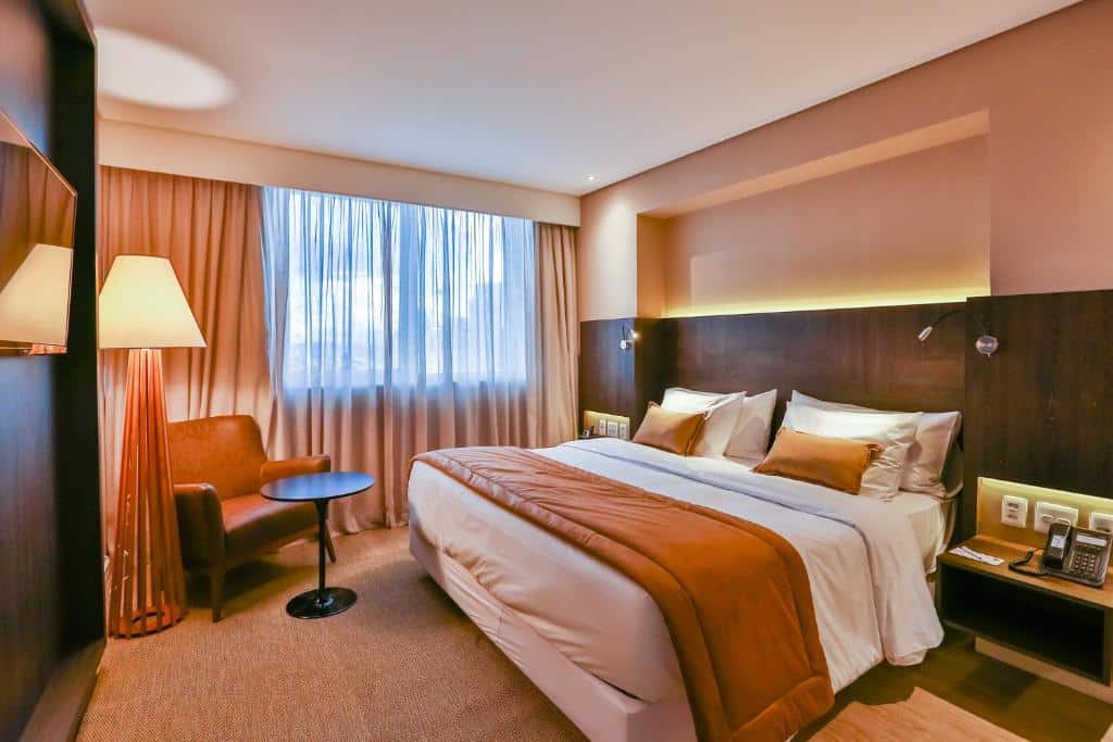 Quarto com cama de casal com colcha branca e marrom, poltrona, pequena mesa a frente, grande abajour, cortinas bege e cabeceira de madeira. Imagem para ilustrar o post hotéis em Goiânia. 