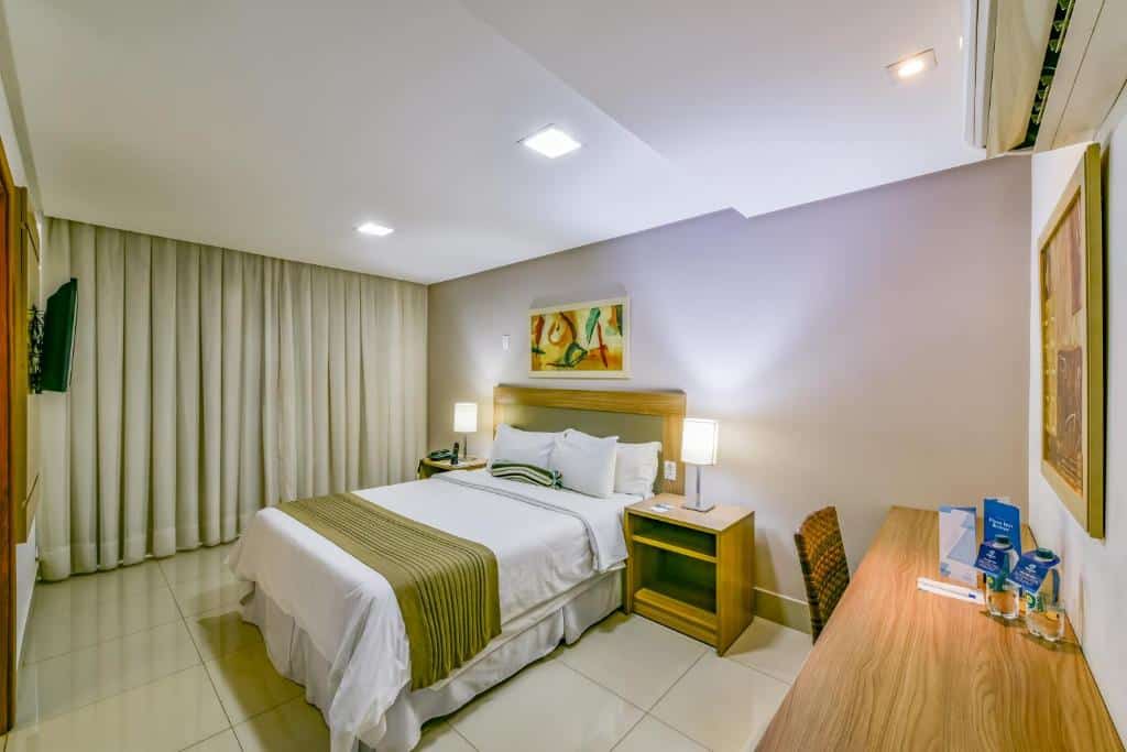 Quarto com paredes e cortinas brancas, cama de casal centralizada com abajour dos lados e quadro decorativo a cima e do lado direito do quarto fica uma mesa com cadeira de madeira. Imagem para ilustrar o post hotéis em Goiânia.