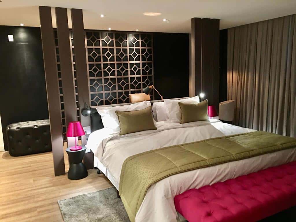 Quarto com cama de casal grande com colcha branca e marrom, banco comprido rosa a frente da cama, abajour rosa ao lado da cama, cortina marrom do lado direito do quarto e paredes pretas. Imagem para ilustrar o post hotéis em Goiânia.