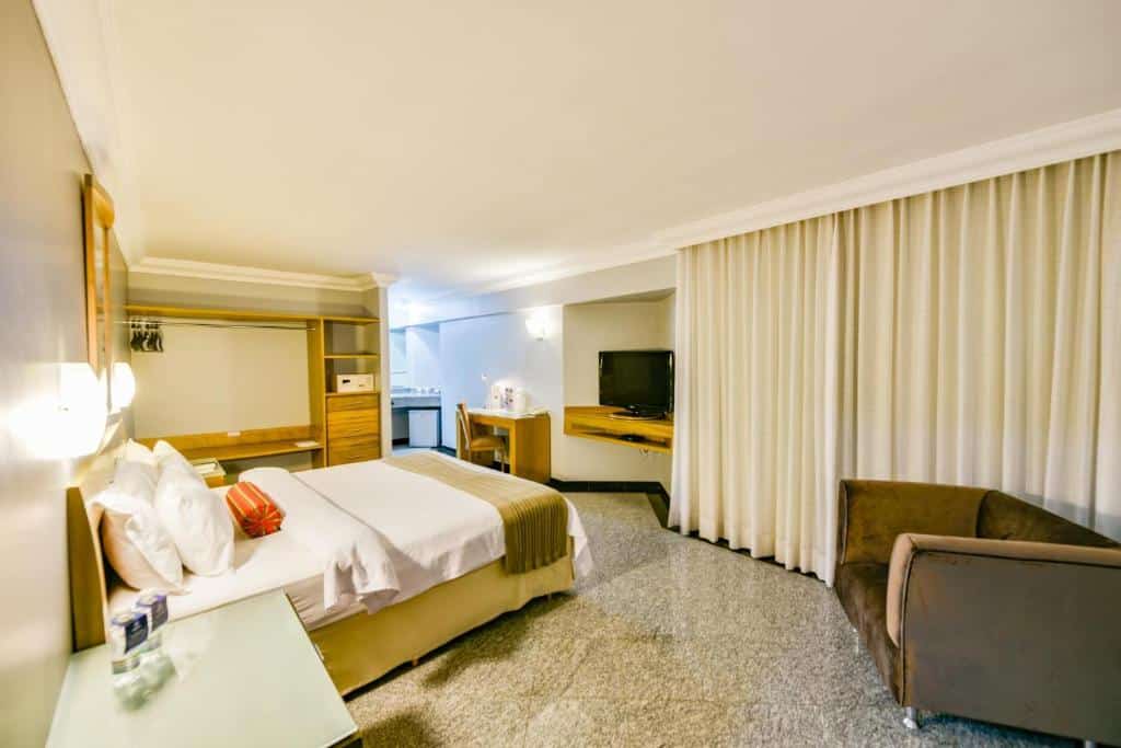 Quarto espaçoso com cama de casal no meio, poltrona cinza no lado direito, armário de maderia no lado esquerdo, cortinas longas bege e TV em frente a cama. Imagem para ilustrar o post hotéis em Goiânia.