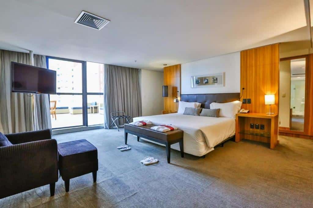 Grande quarto com cama de casal centralizada, paredes brancas, móveis em madeira, poltrona e TV em frente a cama e portas de vidro com longas cortinas que dão para uma varanda. Imagem para ilustrar o post hotéis em Goiânia.