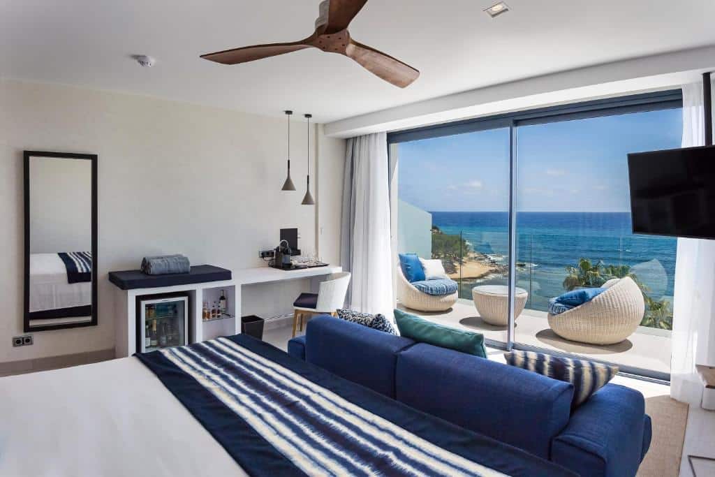 Quarto do Catalonia Royal Ses Estaques, um dos hotéis em Ibiza, moderno com cama, escrivaninha, sofá e varanda com vista para o oceano e duas espreguiçadeiras do lado de fora.