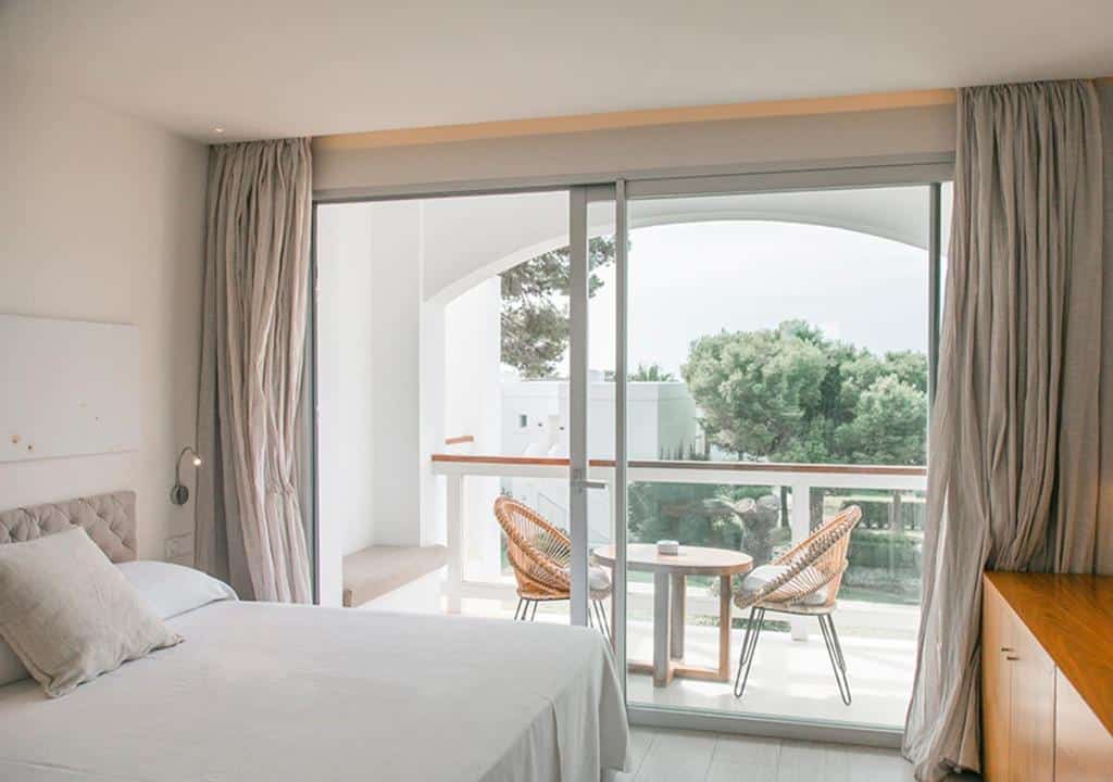 Um quarto do Destino Pacha Ibiza bem iluminado com uma cama, cortinas claras e uma varanda com duas cadeiras e uma pequena mesa com vista para a vegetação.