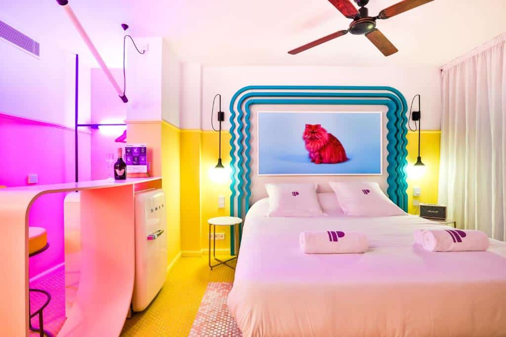 Um quarto de um dos hotéis em Ibiza, o Paradiso Ibiza Art Hotel bem iluminado com uma cama de casal, detalhes em neon, um ventilador de teto e uma foto emoldurada de um gato vermelho acima da cama.