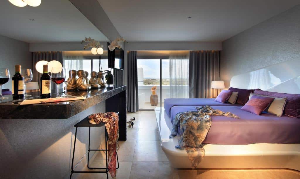 Quarto moderno do Ushuaia Ibiza Beach Hotel com uma cama grande, um balcão de bar, garrafas de vinho e uma varanda com vista para o exterior.