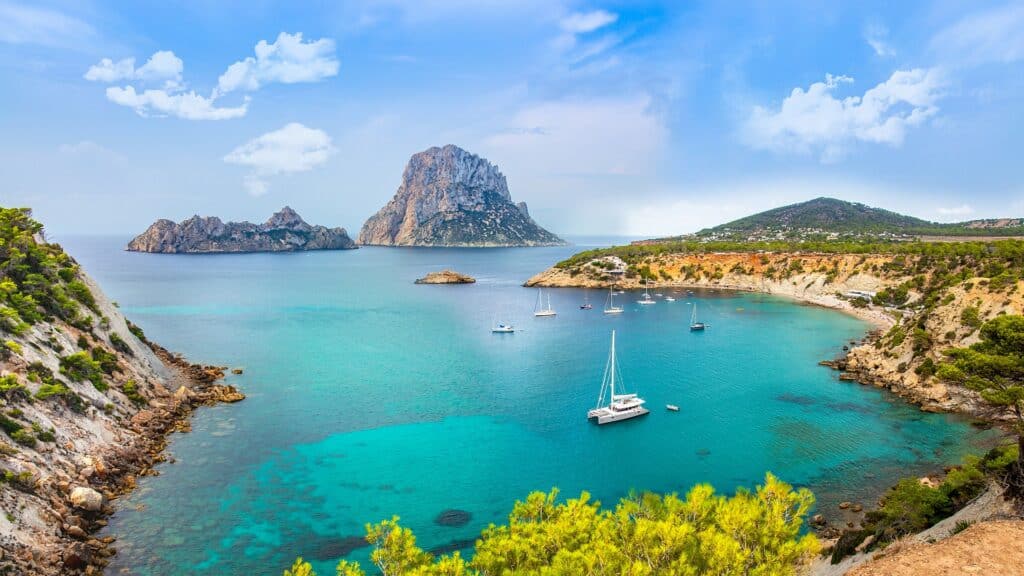 Uma baía azul-turquesa em Ibiza com veleiros ancorados, litoral rochoso, colinas verdes e uma grande ilha ao fundo sob um céu azul.