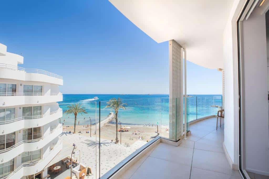 A varanda do One Ibiza Suites um dos hotéis em Ibiza tem vista para uma praia de areia, palmeiras e um mar azul sob um céu claro.