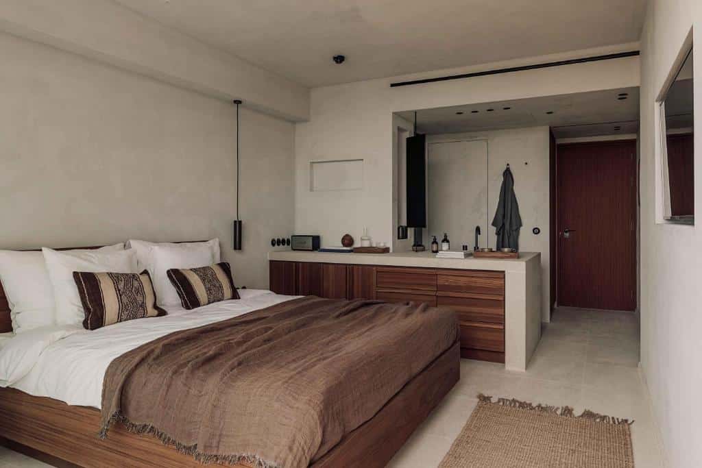 Quarto minimalista do OKU Ibiza com uma colcha marrom, móveis de madeira, paredes brancas e uma porta que leva a uma pequena entrada.