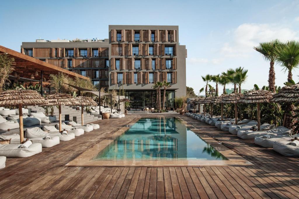 Vista do OKU Ibiza em estilo moderno com detalhes em madeira e vista para uma piscina externa retangular, cercada por espreguiçadeiras e guarda-sóis de palha. Sendo um dos hotéis em Ibiza.
