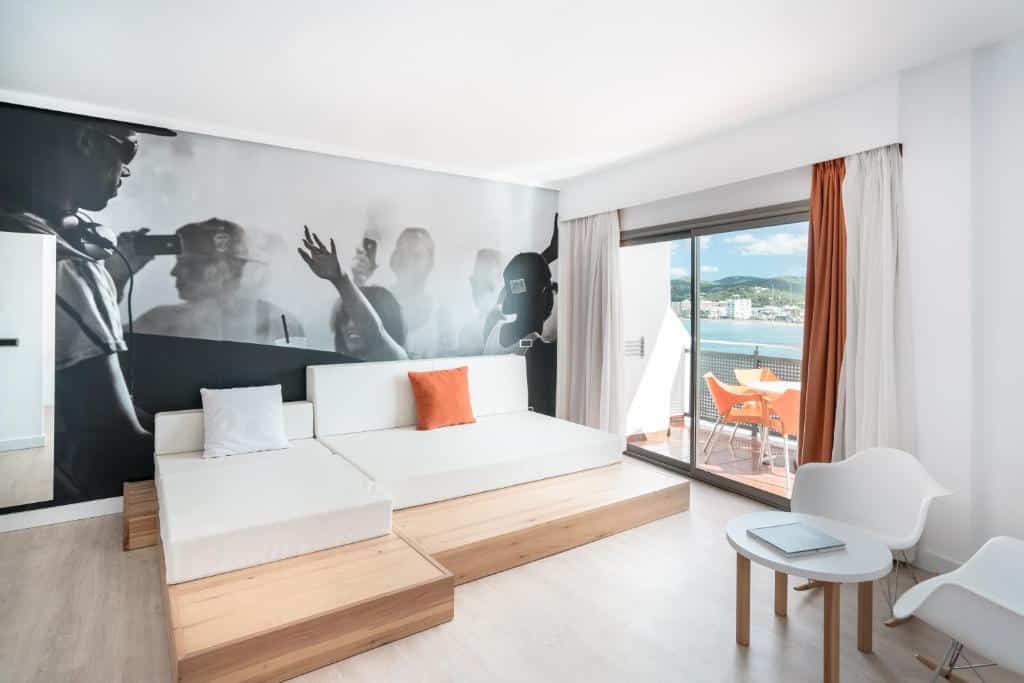 Quarto moderno do THB Ocean Beach com móveis brancos, um mural em preto e branco e uma varanda com vista para a cidade e para a montanha.