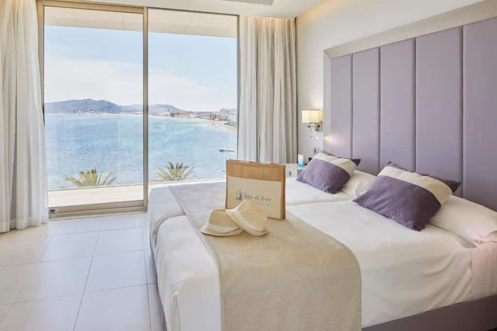 Um quarto do Hotel Torre del Mar com camas de solteiro, cabeceira roxa, vista para o mar através de grandes janelas e um saco de papel com chapéus sobre a cama.