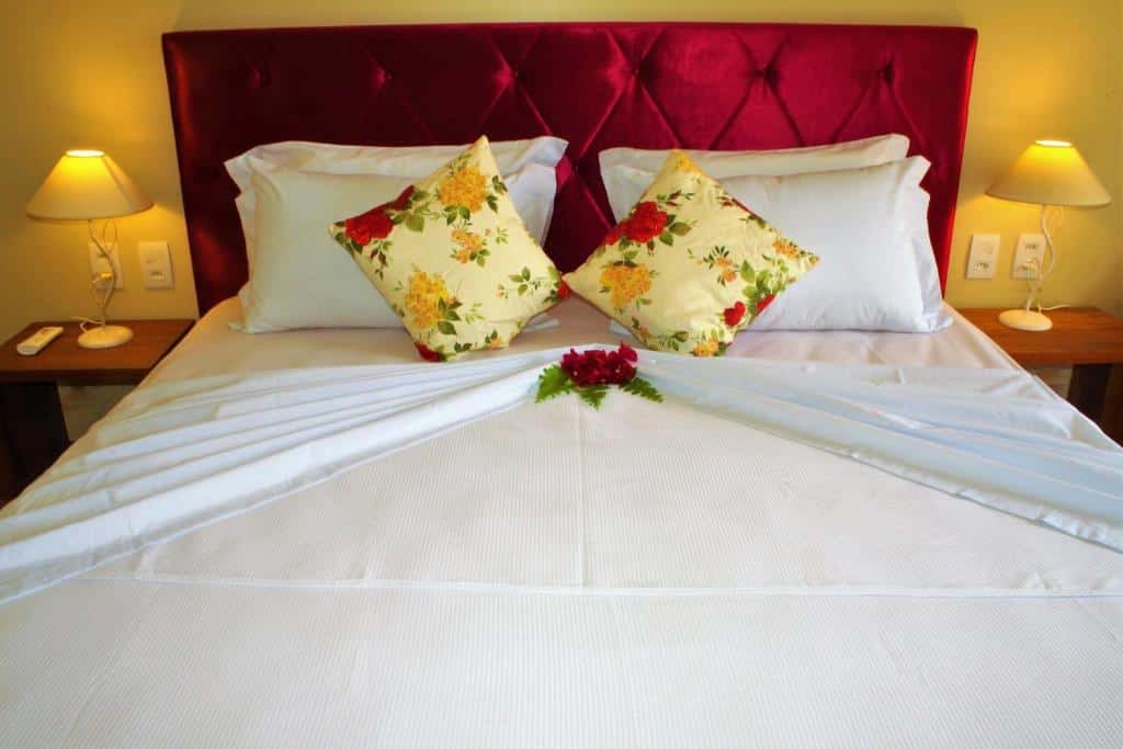 Uma cama feita com lençóis brancos, travesseiros florais, cabeceira vermelha, duas lâmpadas de cabeceira e um arranjo de flores sobre a cama.