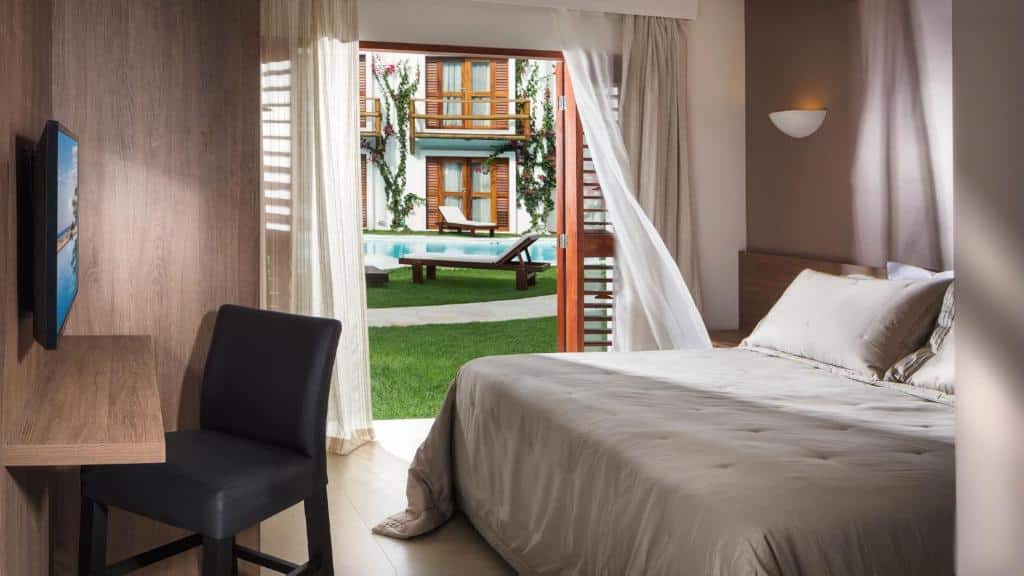 Um quarto de hotel com cama e escrivaninha, portas abertas que dão para um jardim com espreguiçadeiras e uma piscina externa.