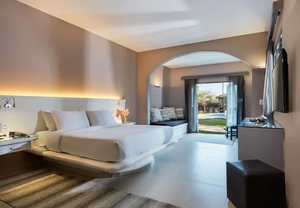 Quarto de hotel moderno com uma cama grande, área de estar, TV e portas abertas com vista para o jardim externo.