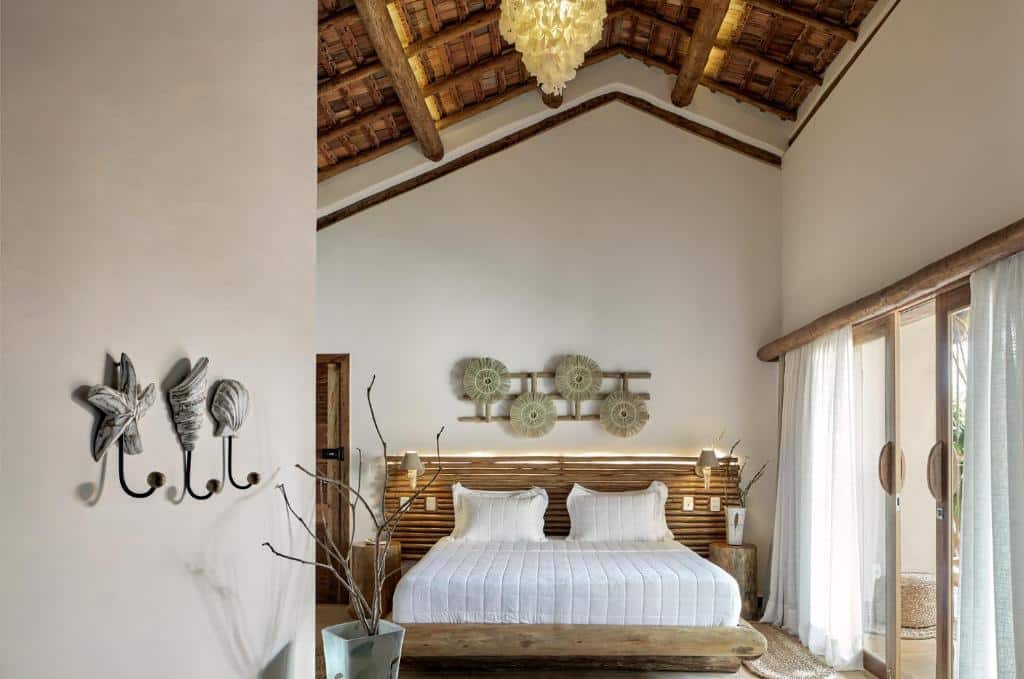 Um quarto com uma cama de madeira, roupa de cama branca, arte decorativa na parede e um teto de palha, com luz solar através de portas de vidro.