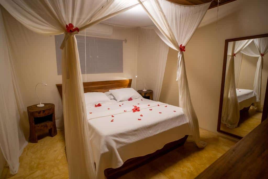 Uma cama de dossel com dossel branco e pétalas de rosas espalhadas, mesas de cabeceira e um grande espelho de parede em um quarto suavemente iluminado.