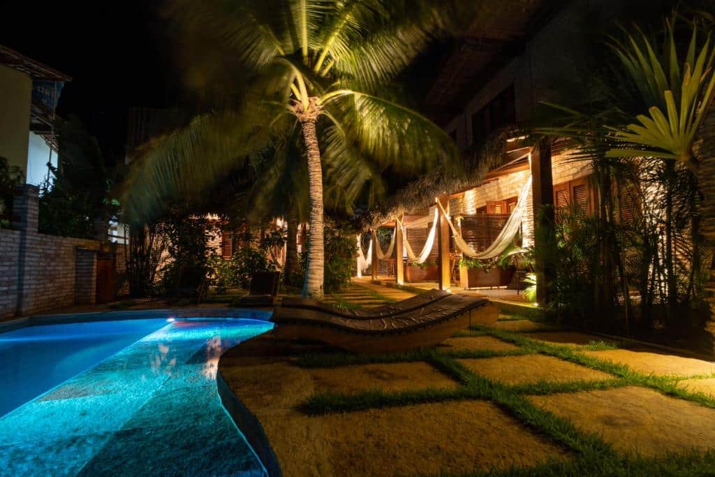 Um quintal à noite com uma piscina iluminada, palmeiras, redes, espreguiçadeiras e um prédio iluminado. Este é um dos hotéis em Jericoacoara.