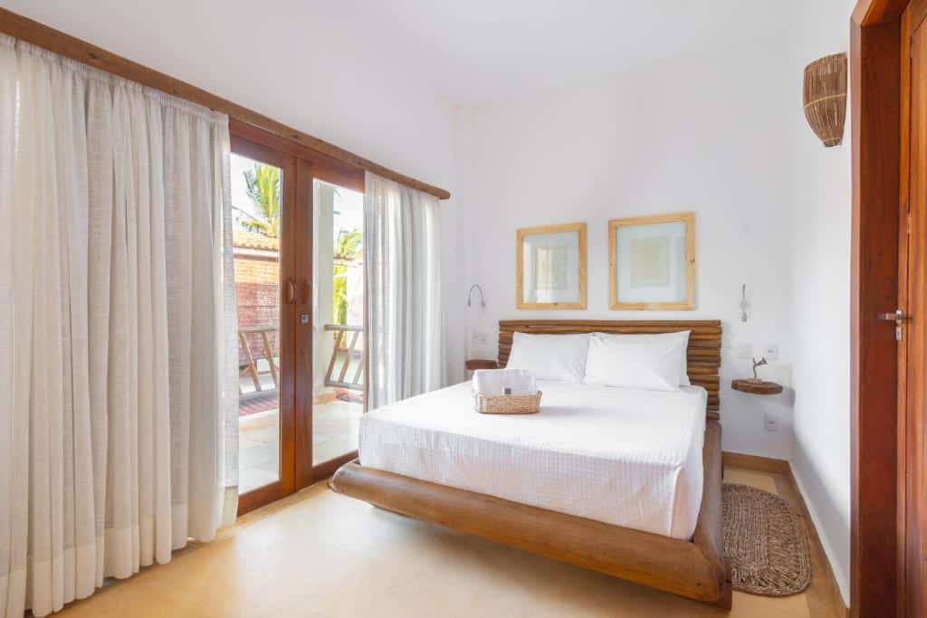 Um quarto bem iluminado com uma cama de madeira, roupa de cama branca, dois quadros emoldurados e portas de vidro deslizantes com cortinas transparentes.
