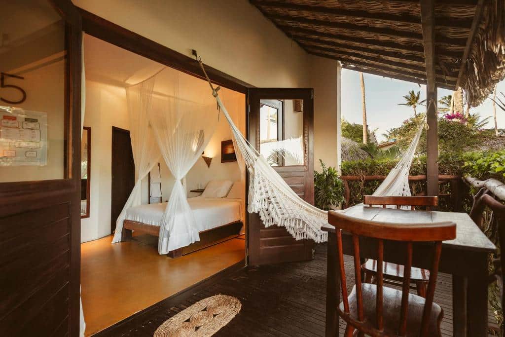 Um quarto de hotel aconchegante com cama e mosquiteiro, que se abre para uma varanda com rede, cadeira e mesa.