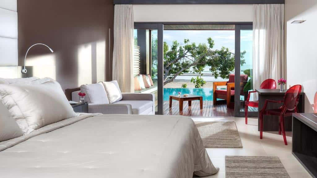 Um quarto de hotel moderno com uma cama grande, área de estar, escrivaninha e portas de correr que se abrem para um pátio com vista para a piscina e o mar.