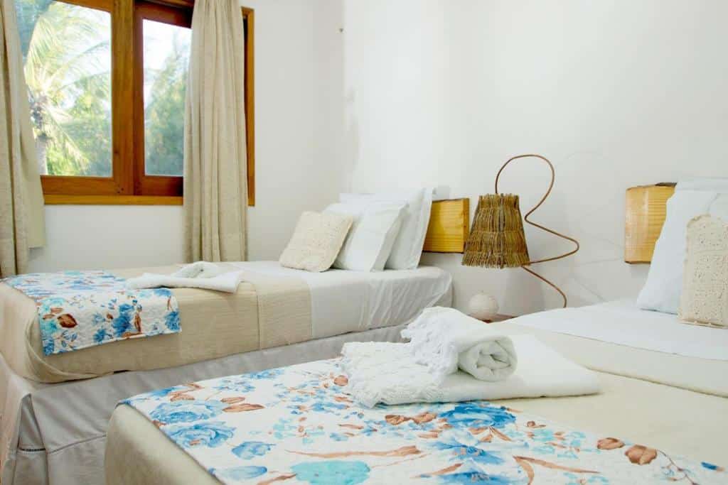 Duas camas de solteiro com roupa de cama floral em um quarto claro, uma janela com cortinas e um abajur em uma mesa de cabeceira entre elas.