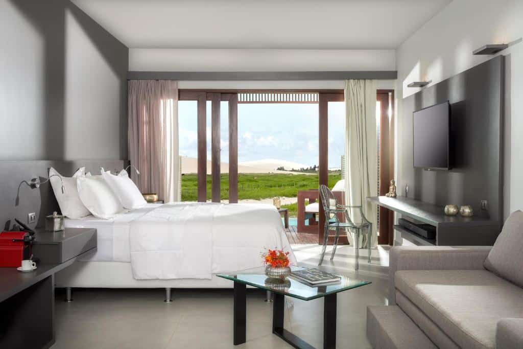 Quarto de hotel moderno com uma cama, sofá, mesa de vidro, TV e janelas grandes com vista para uma área externa gramada.
