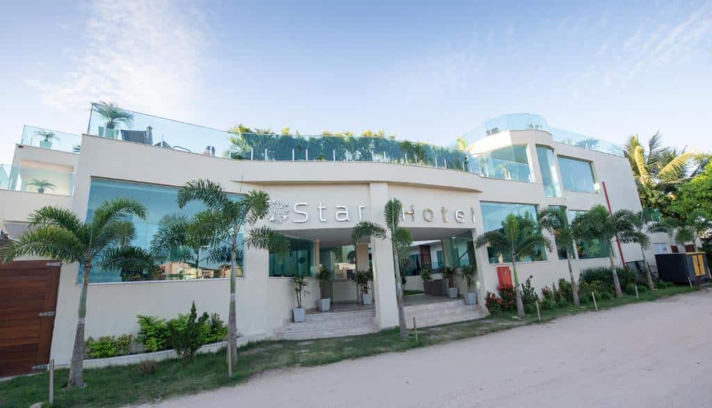 Edifício moderno de hotel com varandas de vidro, palmeiras e uma estrada de areia em frente, sob um céu azul. Este é um dos hotéis em Jericoacoara.