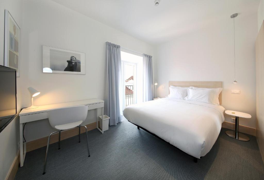 Quarto do Hotel Convento do Salvador com uma cama de casal, ao lado dela há uma pequena sacada com cortinas, mais para frente há uma mesa de escritório com uma cadeira e uma televisão, para representar hotéis em Lisboa.
