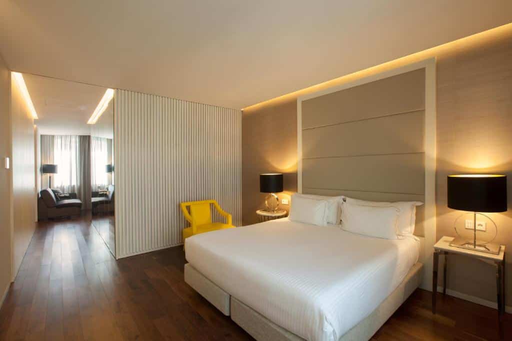 Quarto moderno no Bessa Hotel Liberdade com uma cama grande, cadeira amarela, duas lâmpadas pretas, piso de madeira e uma área de estar ao fundo, para representar hotéis em Lisboa.