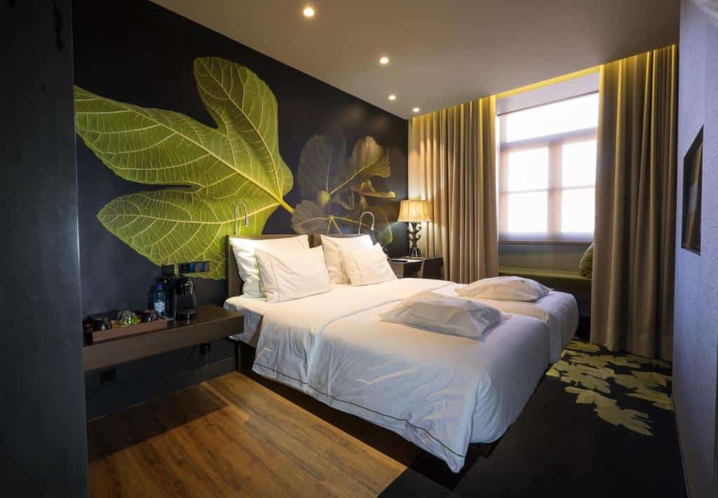 Quarto do Figueira by The Beautique Hotels com uma cama de casal, uma janela com cortinas, há do lado esquerdo da cama uma bancada com itens de café da manhã, e do lado direito há uma pequena mesa com uma cadeira, para representar hotéis em Lisboa.