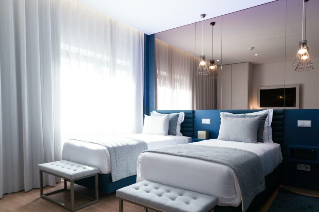Um quarto moderno no Zurin Charm Hotel com duas camas de solteiro, bancos cinza ao pé, luzes penduradas e janelas grandes com cortinas transparentes, para representar hotéis em Lisboa.