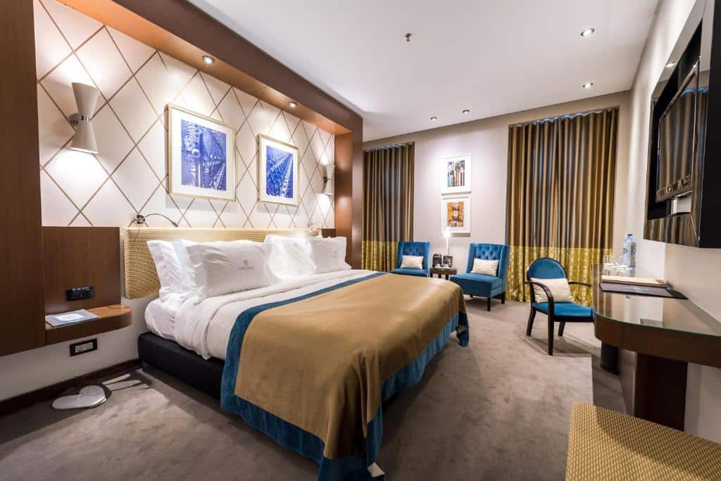Quarto moderno no Lisboa Carmo Hotel com uma cama grande, duas cadeiras, uma escrivaninha e arte na parede, com toques neutros e azuis, para representar hotéis em Lisboa.