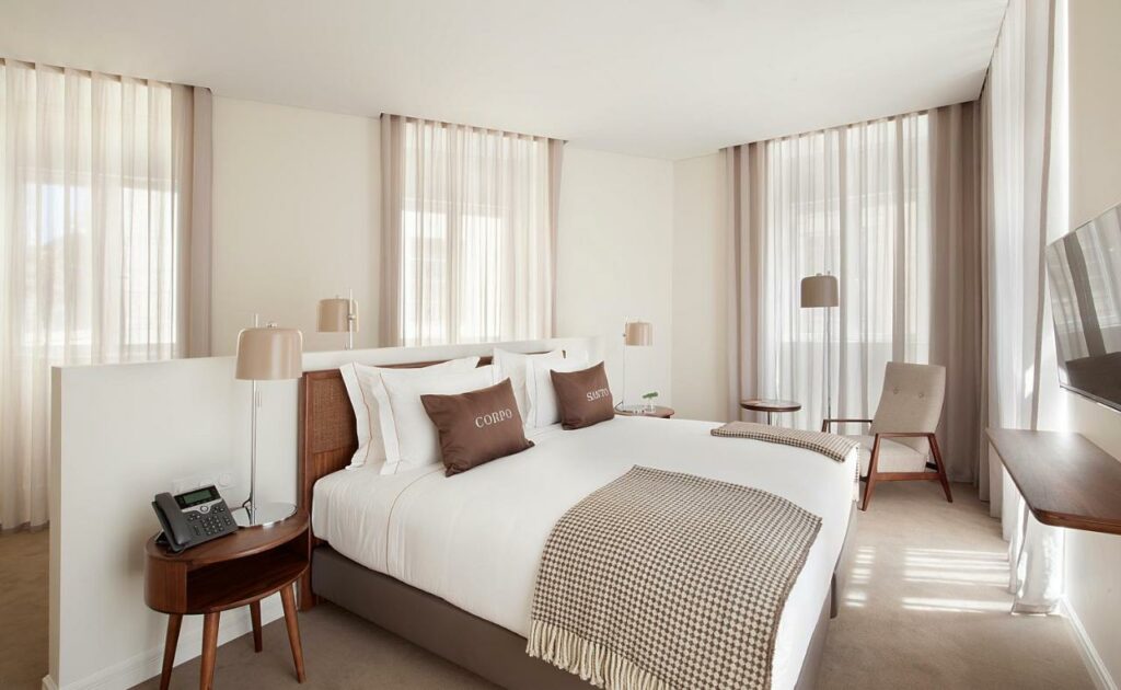 Um quarto moderno no Corpo Santo Lisbon Historical Hotel com uma cama grande, decoração neutra, duas cadeiras, uma mesa lateral e uma TV na parede, para representar hotéis em Lisboa.