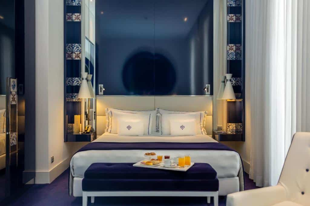 Quarto do Portugal Boutique Hotel com uma cama de casal, uma janela com cortinas, uma poltrona branca, um banco ao pé da cama com uma bandeja com café da manhã, para representar hotéis em Lisboa.