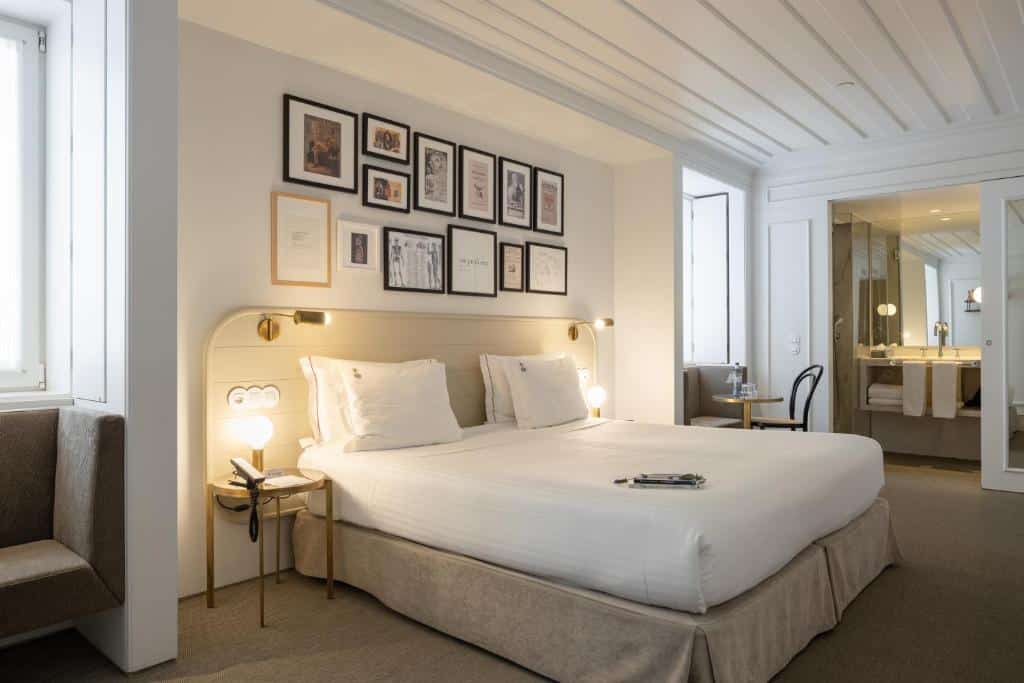 Quarto do Lisboa Pessoa Hotel com uma cama de casal, na parede atrás da cama há diversos quadros, nas laterais da mesma há mesinhas com luminárias, há duas janelas do cômodo, para representar hotéis em Lisboa.