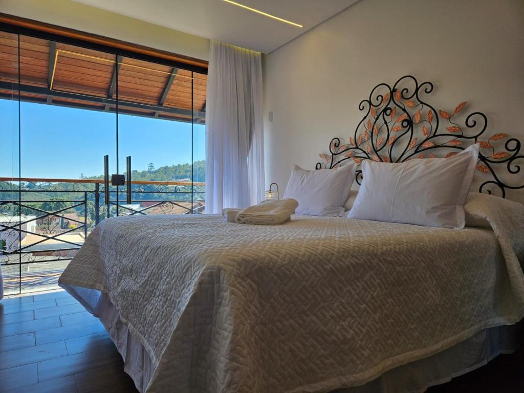 Uma cama bem arrumada com roupas de cama e toalhas brancas está em um quarto bem iluminado com uma grande janela com vista para a paisagem, no Hotel Villa Greenberg, para representar hotéis em Monte Verde.