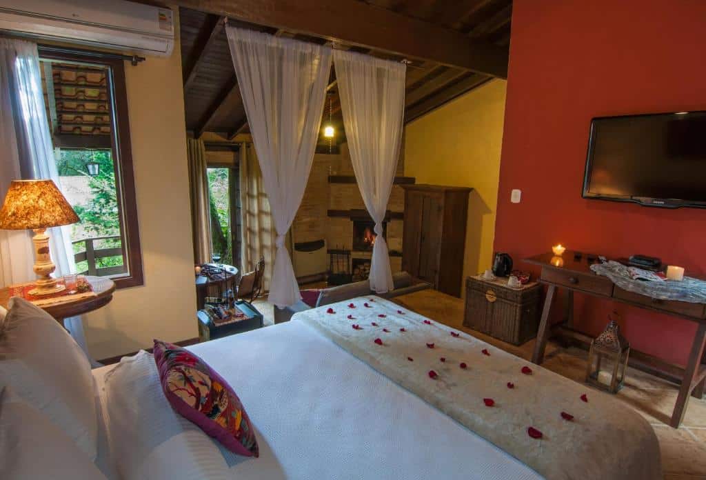 Um quarto aconchegante com uma cama de casal, pétalas de rosa, iluminação aconchegante, uma TV e uma área de estar com janelas com cortinas, na Estalagem Wiesbaden, para representar hotéis em Monte Verde.
