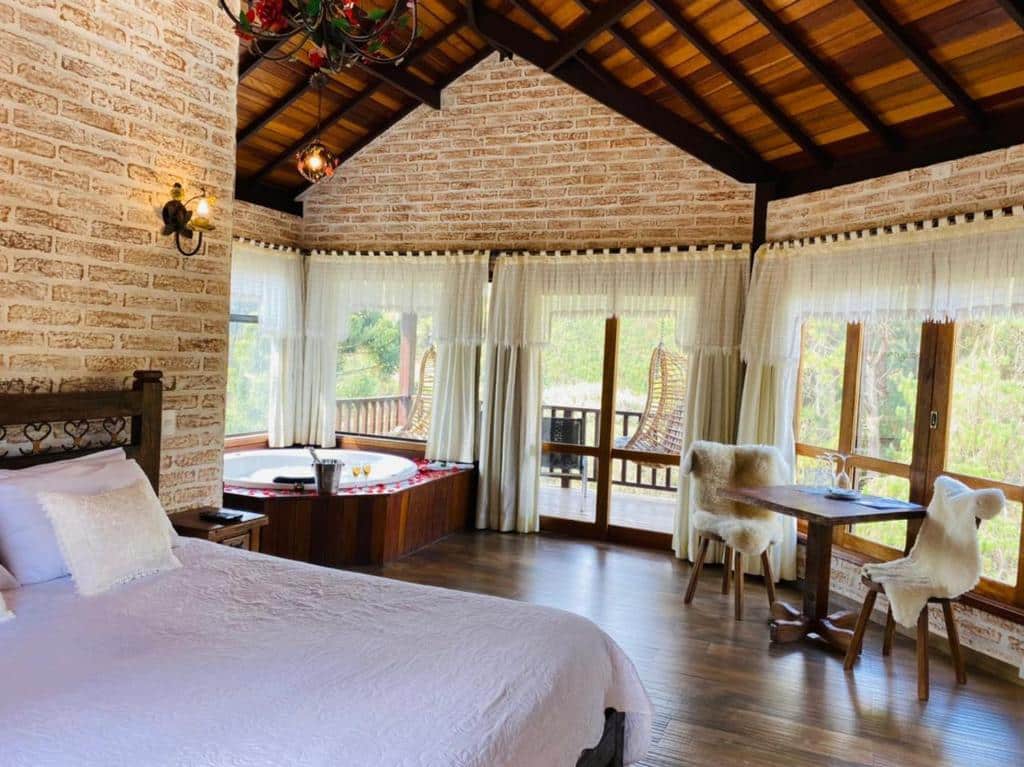 Um quarto aconchegante na Estalagem Mandeville com uma cama grande, uma banheira de hidromassagem ao lado da janela, uma pequena mesa com cadeiras e janelas grandes que permitem a entrada de luz natural, para representar hotéis em Monte Verde.