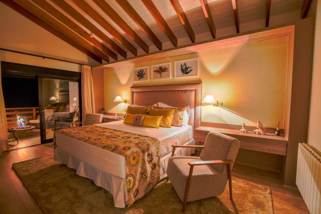 Um quarto com iluminação aconchegante no Hotel Saint Michel, com uma cama grande, roupa de cama estampada, poltrona, vigas de madeira e arte na parede acima da cabeceira, para representar hotéis em Monte Verde.